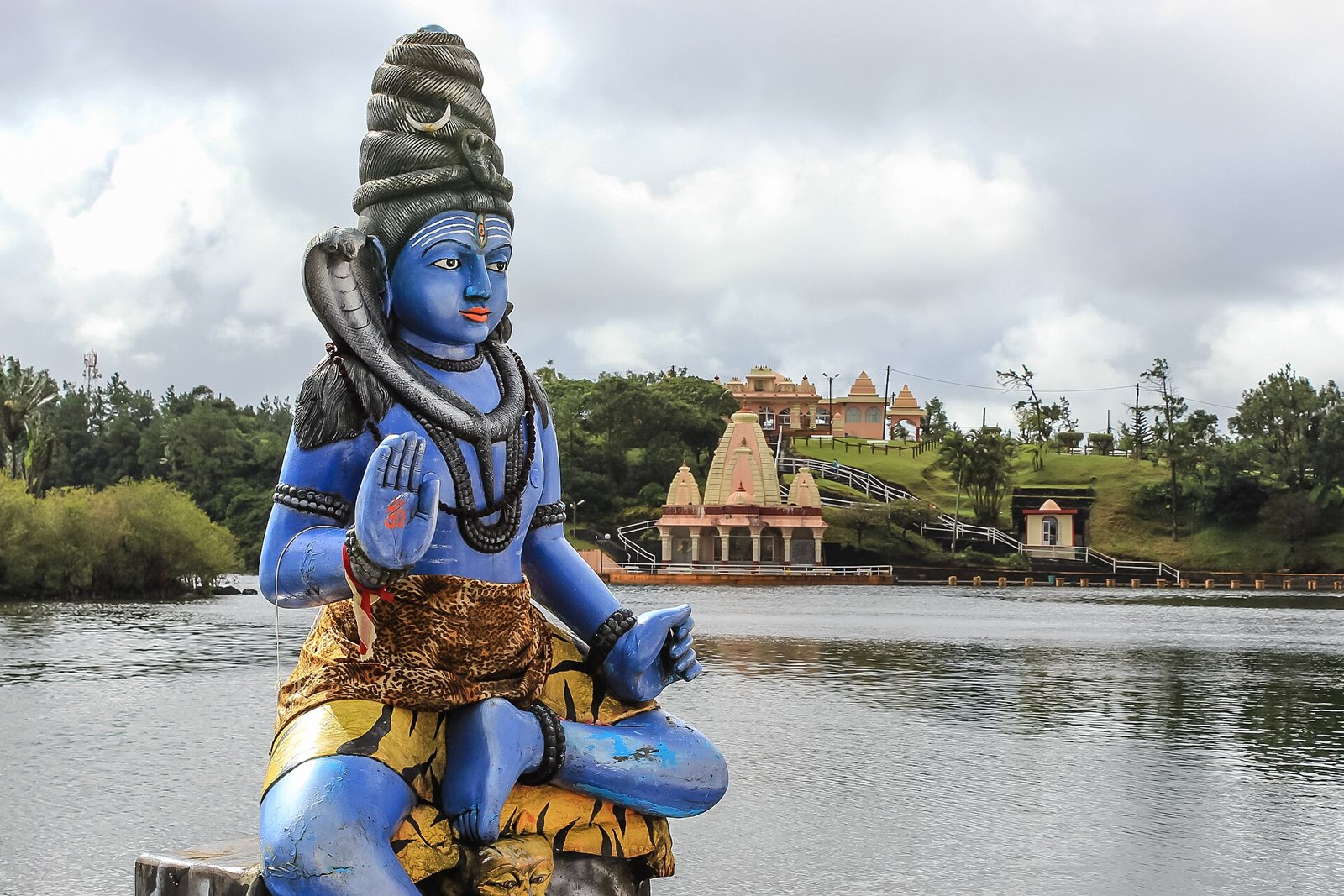 Blaue Shiva-Statue am Seeufer, dahinter ein Tempel